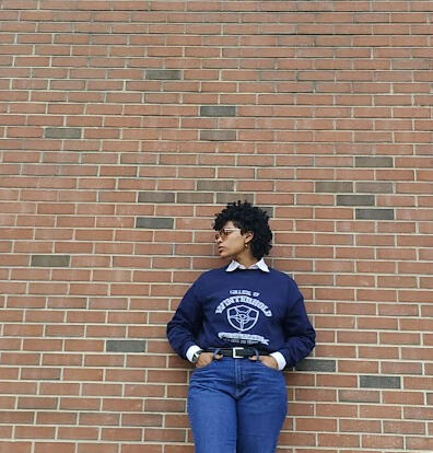 A photo of Ares wearing an all blue academic-ish outfit in front of a brick background as they look to the side.